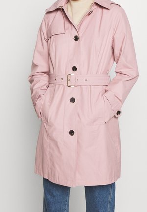 Kvinde iført en lyserød trenchcoat med bælte og sorte knapper, hænderne i lommerne, kombineret med blå jeans foran en ensfarvet baggrund.