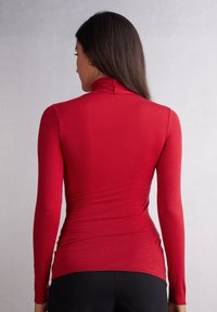 Rotes Langarmoberteil mit hohem Kragen, das eine figurnahe Silhouette und eine glatte Textur aufweist. Die Rückseite zeigt Nähte und einen nahtlosen Übergang am Kragen.