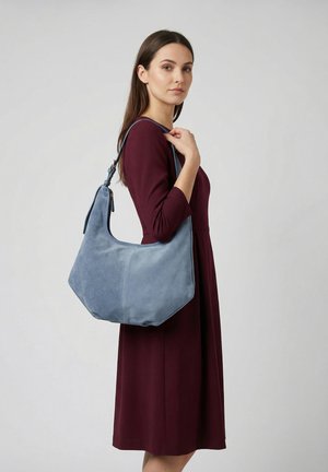 Vrouw in een bordeauxrode jurk met een grote lichtblauwe suède schoudertas aan haar rechterschouder tegen een effen lichte achtergrond.