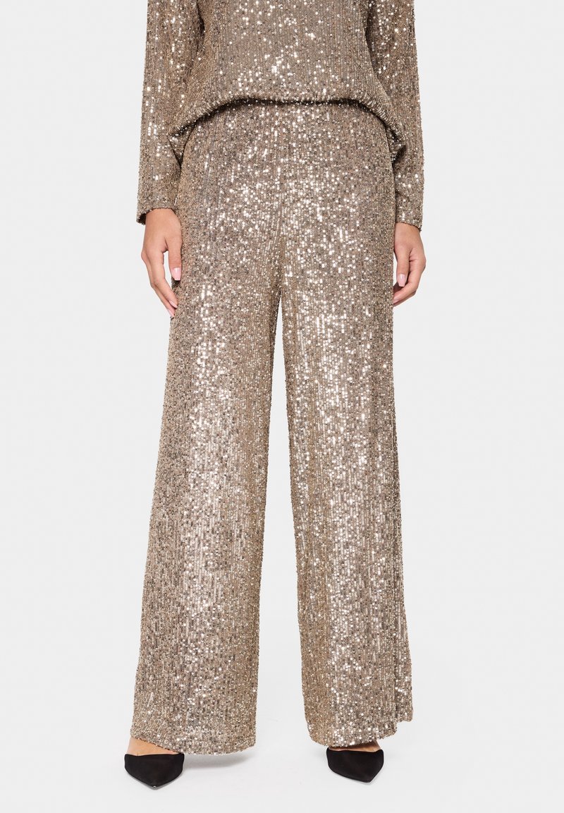 Ensemble deux pièces en sequins dorés avec des manches longues et un pantalon à jambes larges. Texture lisse avec des détails réfléchissants et scintillants ; chaussures noires.