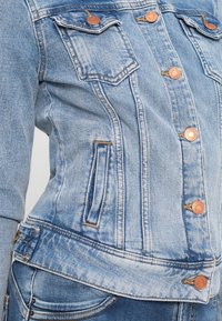 Svetlo modra jeans jakna s prilegajočim dizajnom, gumbom za zapiranje, dvema žepoma na prsih in kontrastnim šivom ob šivih.