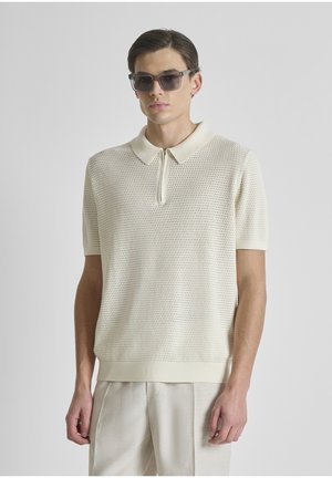 Joven con gafas de sol rectangulares oscuras, polo de punto crema de manga corta con cuello con cremallera y pantalones de color claro, sobre un fondo liso.