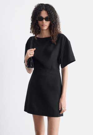 Femme aux cheveux bouclés portant des lunettes de soleil noires et une robe noire à manches courtes, tenant un petit sac noir sur l'épaule.