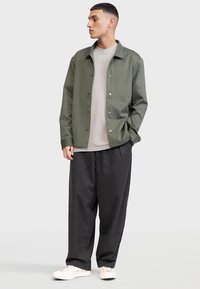 Olive roheline jakk pehme tekstuuriga, nööpidega kinni, kantud heledast hallist T-särgist ja tumedatest triibulistest pükstest, komplekteeritud valgete kingadega.