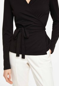 Zwarte wikkeltop met lange mouwen, voorzien van een strik op de taille. Glad materiaal met een V-hals ontwerp, gecombineerd met een witte broek.