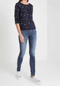 Felpa navy con motivo floreale e maniche lunghe, abbinata a jeans skinny blu e sneakers argentate, ambientata su uno sfondo chiaro.