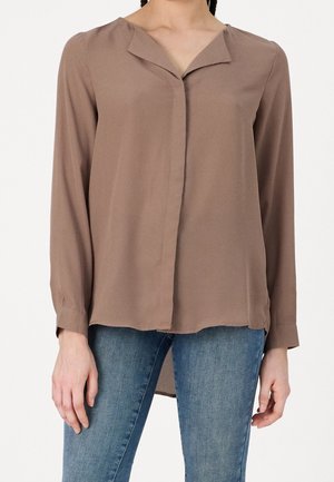 Woman wearing a loose, long-sleeve brown blouse with a folded collar paired with blue denim jeans.