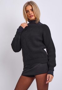 Schwarzer Strickpullover mit hohem Kragen, der mit strukturierten Mustern und gerippten Bündchen versehen ist, kombiniert mit einem schwarzen Minirock und transparenten Strumpfhosen.