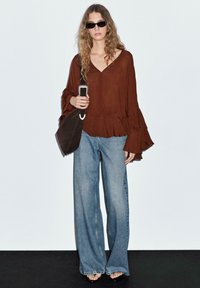 Maglione marrone a maniche lunghe con polsini arricciati; jeans in denim blu a gamba larga; occhiali da sole rettangolari neri; tiene una borsa scura con una fibbia argentata.