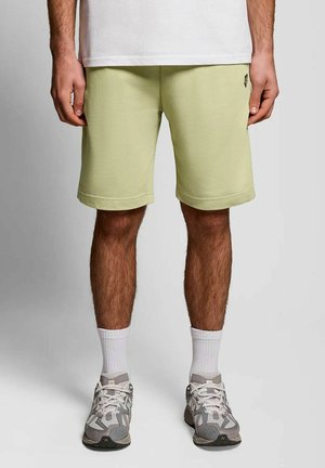 Mann trägt hellgrüne knielange Shorts, weiße Socken und graue Sportschuhe und steht vor einem einfarbigen Hintergrund.