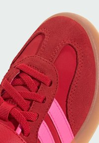 Röd sportskor med överdel i mocka och textil, rosa ränder och rund sula. Snören i matchande röd säkrar passformen.