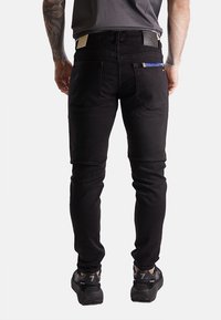 Jeans neri slim-fit con tasche posteriori, etichetta in pelle color cuoio e un accento blu visibile sulla tasca destra. Progettati con una texture liscia.