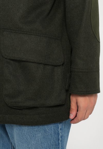 Dark green wool coat with a smooth texture, featuring a large side pocket and a tailored fit. Complemented by light blue denim.
