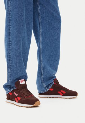 Smeđe antilop tenisice s crvenim detaljima, bijelim srednjim slojem i gumenim potplatom boje tamnog bež. Uparene s plavim trapericama. Logotip na jeziku i bočnoj strani.