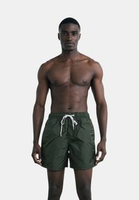 Pantaloni da bagno verdi realizzati in materiale ad asciugatura rapida, con vita elastica e coulisse bianca, indossati da un modello maschile senza maglietta.