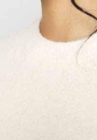 Vit stickad tröja med rund halsringning, som har en strukturerad yta och ribbad kant. Tyget ser mjukt och tjockt ut.