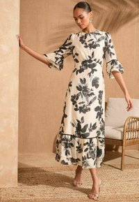 Cvetlična maksi obleka v kremni barvi s črnimi in zeleno-modrimi poudarki, z volanastimi rokavi, večslojnim robom in poudarjenim pasom. Minimalističen dizajn.