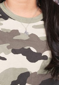 Silberne Halskette mit einem Anhänger in Form eines galoppierenden Pferdes, getragen über einem T-Shirt mit grünem und grauem Tarnmuster. Glatte Textilien mit taillierter Passform.