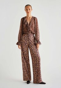 Langärmlige Bluse mit gerafftem Ausschnitt im braunen und lila geometrischen Muster, kombiniert mit weit geschnittenen Hosen im gleichen Design.