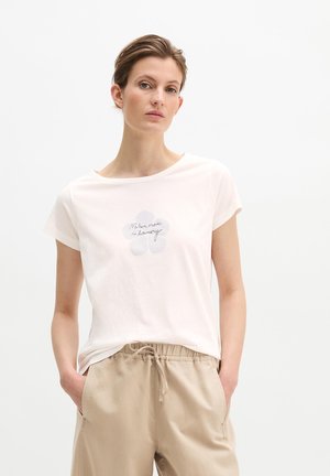 Kobieta w białej koszulce z delikatnym kwiatowym nadrukiem i tekstem pisanym kursywą, w beżowych spodniach z troczkiem, z rękami w kieszeniach, na neutralnym tle.