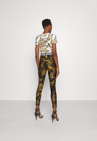 Weißes Top mit goldenen, kunstvollen Mustern, kombiniert mit schwarzen Leggings mit goldenen Designs. Schwarze hochhackige Sandalen mit Knöchelriemen.