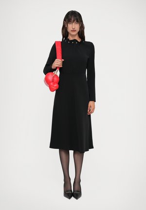 Vestito lungo nero a maniche lunghe con colletto alto e bottoni decorativi, abbinato a una borsa a forma di cuore rossa e tacchi a punta neri.