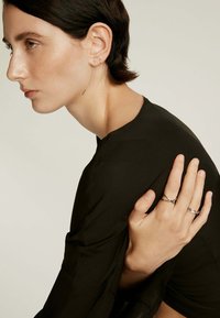 Pendientes de plata con un delicado detalle de cadena, anillos con varios diseños y una blusa negra de manga larga y suave en el perfil de un modelo.
