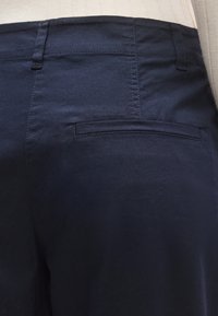 Pantaloni din bumbac albastru marin cu o textură fină, având un buzunar posterior cu clapetă și cusături întărite. Prim-plan pe secțiunea superioară din spate.