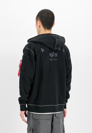 Man draagt een zwarte Alpha Industries hoodie met logo op de rug en grijze cargobroek, staand met zijn rug naar een witte achtergrond.