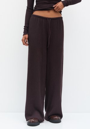 Personne portant un pantalon large texturé marron foncé avec une taille à cordon et des chaussures marron arrondies sur un fond blanc uni.
