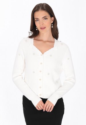 Vrouw draagt een witte gebuttonde cardigan met parelachtige details en geschulpte randen, gecombineerd met een zwarte rok, poserend tegen een witte achtergrond.