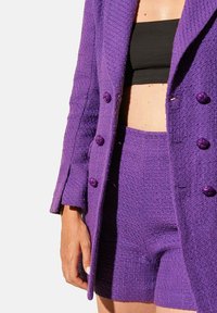 Blazer texturé violet avec shorts assortis ; présente des boutons décoratifs violets et un crop top noir en dessous.
