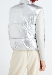 Silberner Puffer-Westen mit hohem Kragen, der eine glänzende, glatte Textur und horizontale Nähte aufweist. Trägt man über einem hellgrauen, langärmeligen Oberteil.