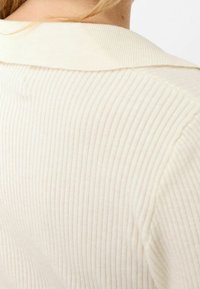 Gros plan sur le dos d'une personne portant un pull en maille côtelée de couleur crème avec un large col plié.