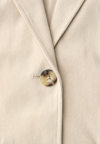 Lähikuva beigevärisestä, teksturoidusta kangastakista, jossa on yksi suuri pyöreä nappi, johon on kaiverrettu teksti ja joka kiinnittää kauluksen.