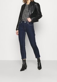 Veste courte en cuir noir, haut à motif floral, jean en denim foncé avec surpiqûres contrastantes, et bottines noires à talons de hauteur cheville.
