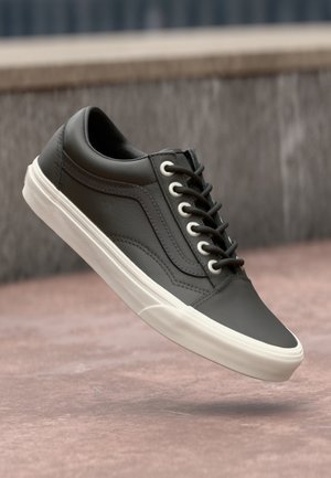 Czarny niski sneaker skórzany z białą podeszwą i czarnymi sznurowadłami unoszący się nad betonową powierzchnią i szarym tłem ściany.