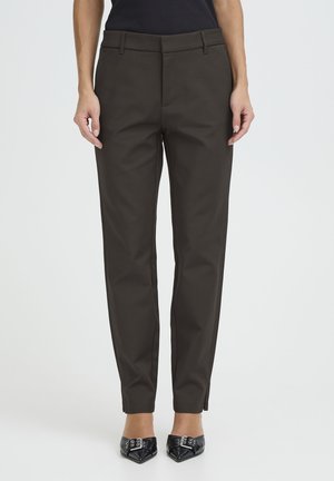 Nő, szűk szabású sötét olívazöld nadrágot és fekete, hegyes orrú magassarkút visel, és egy egyszínű világos háttér előtt áll.