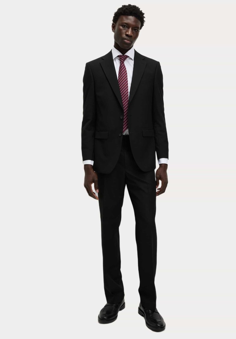Jonge man staat rechtop, gekleed in een zwart pak, wit overhemd, rood gestreepte stropdas en zwarte nette schoenen tegen een effen lichte achtergrond.