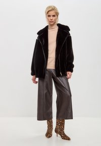 Zwarte nepbont jas met brede revers en ritssluiting, gecombineerd met grijze glanzende wijde broek en enkellaarsjes met luipaardprint.