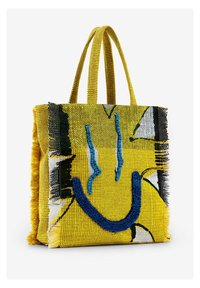 Żółta pleciona torba na ramię z graficznym wzorem uśmiechniętej buźki w czerni i niebieskim. Posiada fakturowane, frędzlowane krawędzie oraz mocne, podwójne uchwyty.