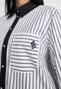 Hvit og svart stripete skjorte med svart krage og lomma. Har metallknapper og en svart brodert logo på lomma.
