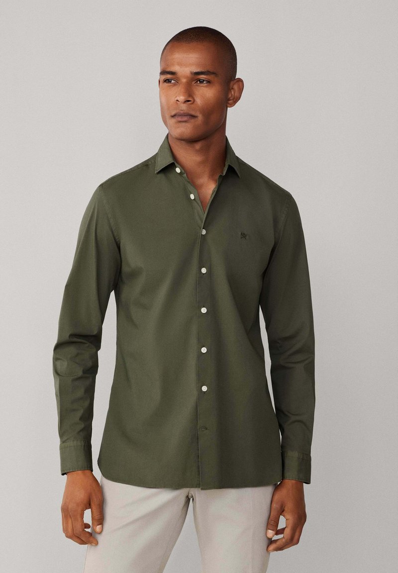 Olijfgroene blouse met knopen van een gladde stof, met een klein logo op de borst, lange mouwen en witte knopen.