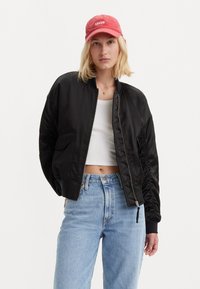 Zwarte satijnen bomberjack met geribbelde manchetten, voorzien van een rits aan de voorkant en zijzakken, gedragen over een wit crop topje en blauwe jeans met hoge taille.