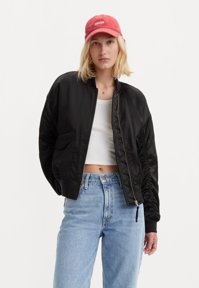 Zwarte satijnen bomberjack met geribbelde manchetten, voorzien van een rits aan de voorkant en zijzakken, gedragen over een wit crop topje en blauwe jeans met hoge taille.