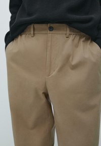 Béžové kalhoty s plochým předním dílem, elastickým pasem a zapínáním na knoflík. Hladká látka s jemným leskem, kombinovaná s černým svetrem.