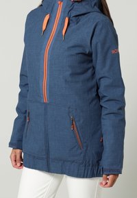 Niebieska kurtka odporna na warunki pogodowe z kapturem, pomarańczowymi zamkami błyskawicznymi i elementami, kieszeniami na zamek, teksturowaną tkaniną oraz regulowanymi mankietami.