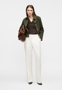 Γυναίκα που φοράει ελιά πράσινο μπουφάν, σκούρο καφέ τοπ, λευκό παντελόνι με φαρδιά πόδια, κρατώντας μεγάλη καφέ δερμάτινη τσάντα, στέκεται σε λευκό φόντο.