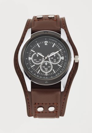 Montre analogique avec cadran noir, trois sous-cadrans, boîtier argenté et large bracelet en cuir marron avec surpiqûres orange et rivets métalliques.