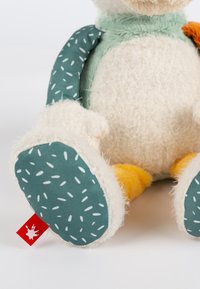 Soft plush toy with cream body, teal patterned arms, and yellow feet. Features red label on the side. Textured for tactile engagement.